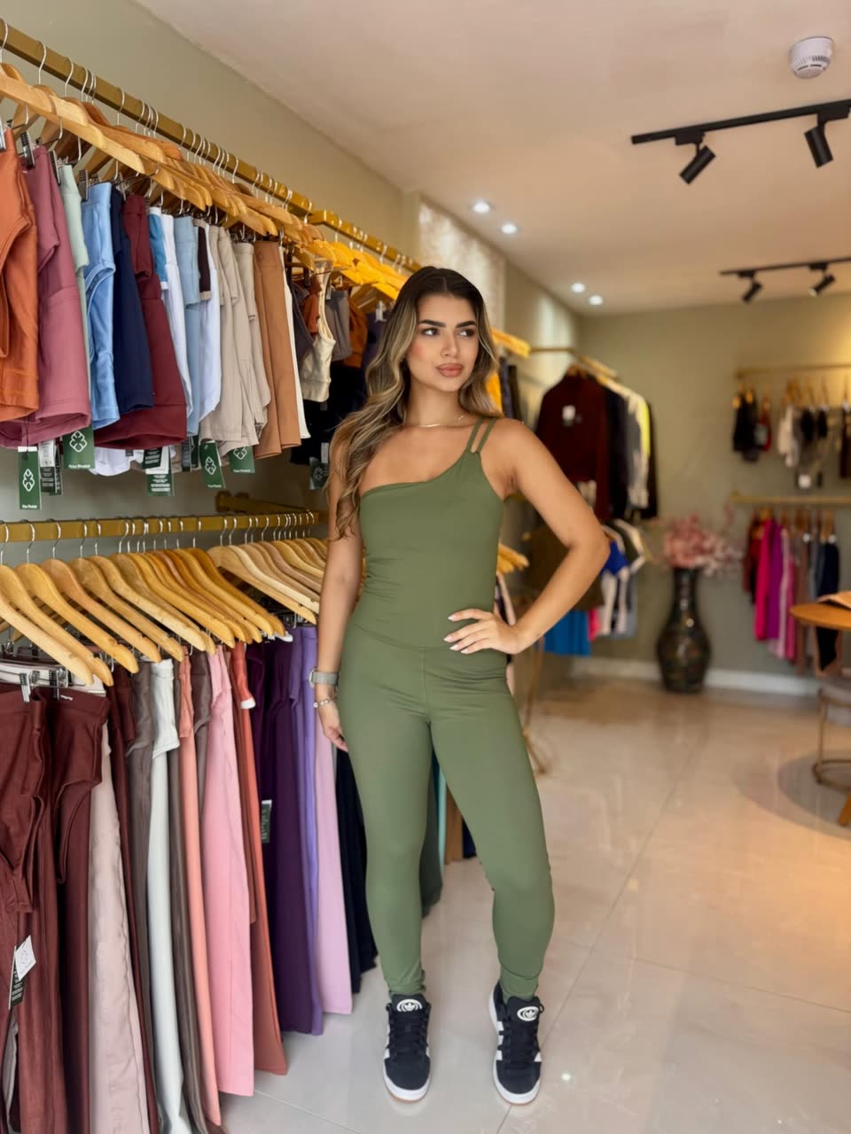 Cliente vestindo o macacão verde oliva dentro da loja Flow Fitwear, com araras de conjuntos coloridos ao fundo